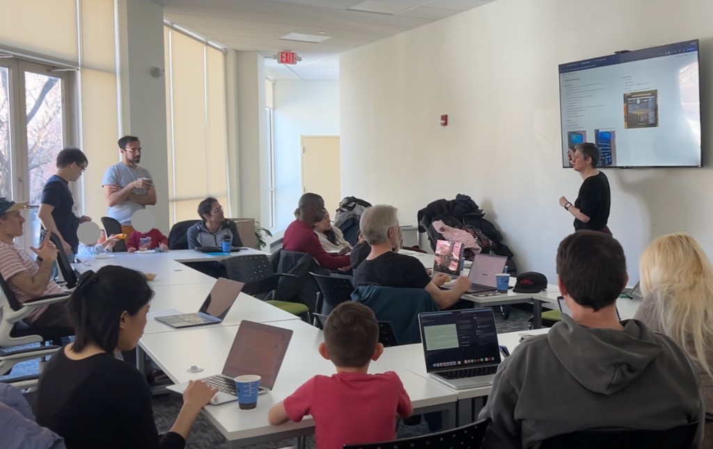 A meetup room where parents and kids sit together at tables with laptops, watching a member present at the front.