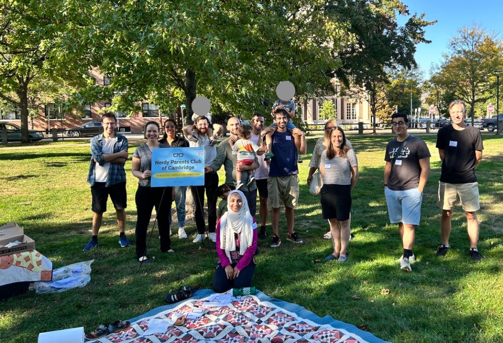 Club members and their kids gathered around a picnic blanket in a park, holding a Nerdy Parents Club of Cambridge sign.