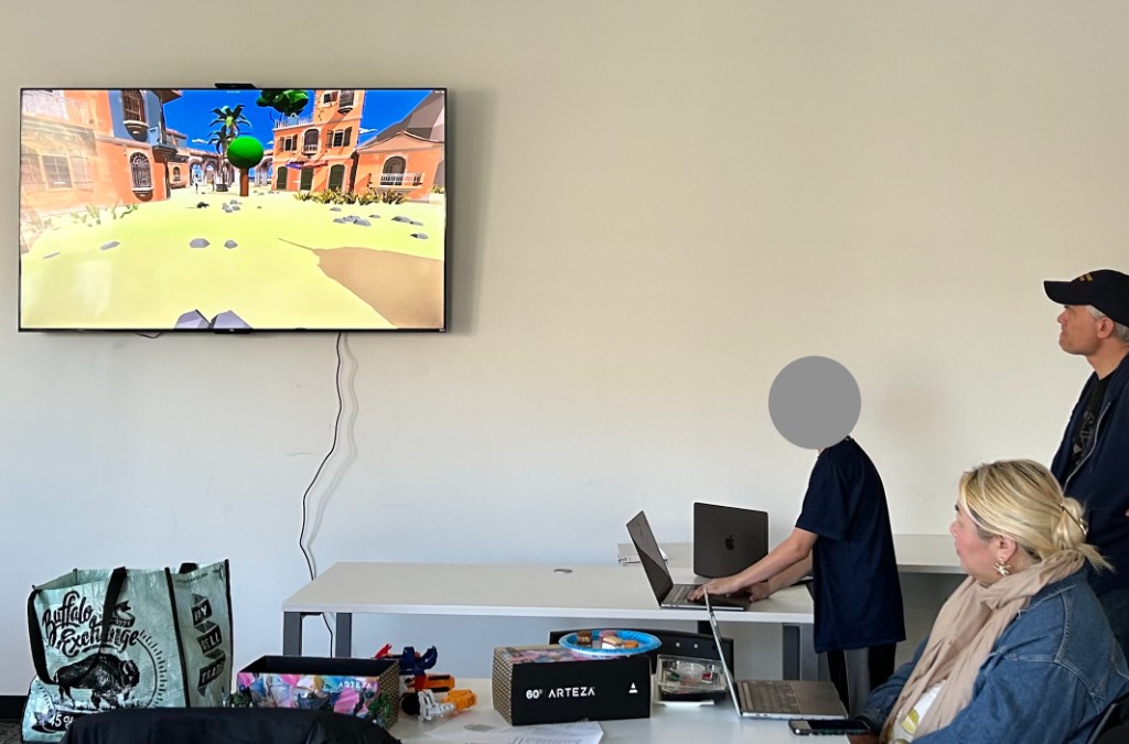 A child standing at a laptop demoing a 3D video game they built, shown on a wall-mounted screen, while other members watch.